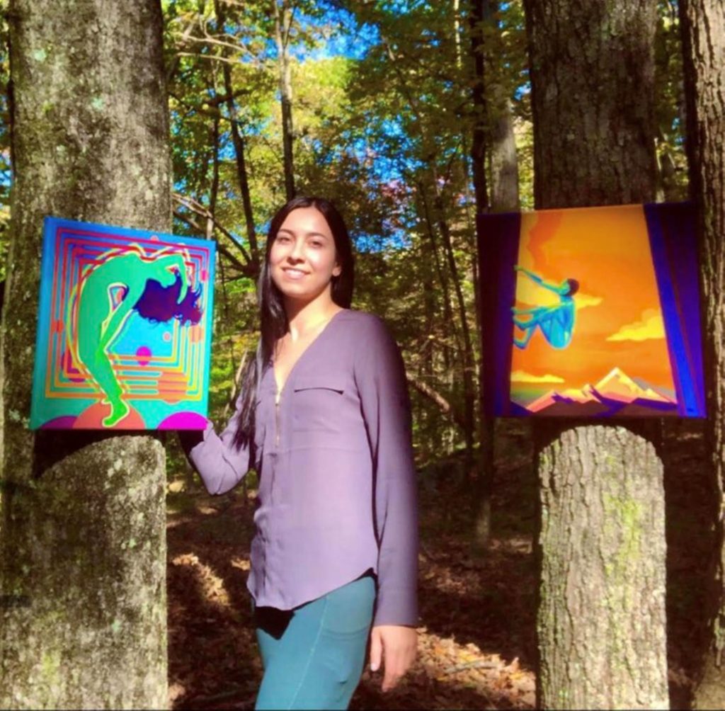 A photograph of artist Cynthia Celone standing in the woods with two of her artworks hanging from trees